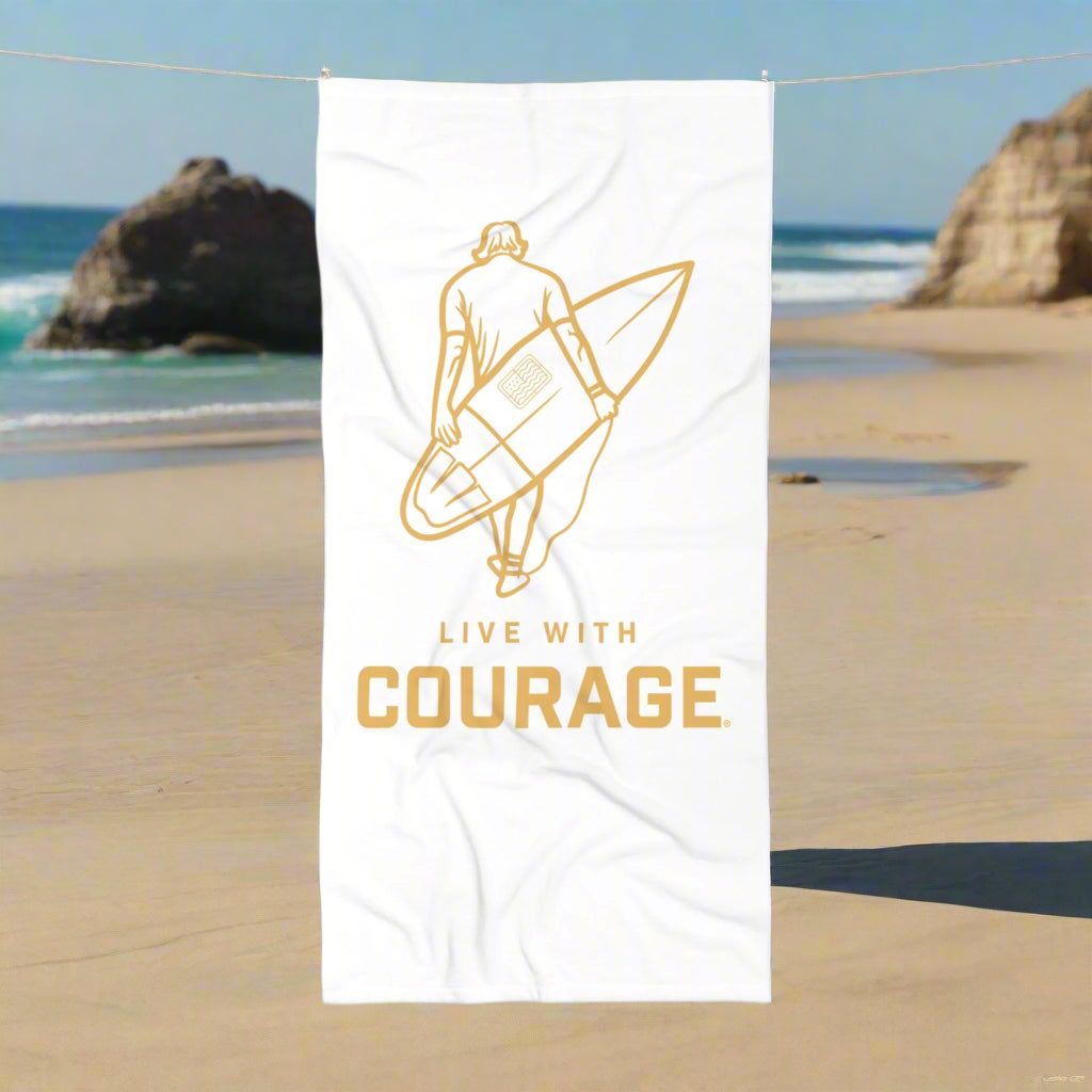 White beach towel with a graphic in gold of a male surfer holding his surfboard on his back and the logo Live With Courage®️ underneath.