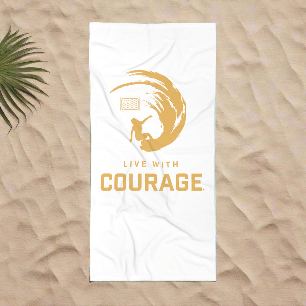 A beach scene with a white 30 by 60 inch beach towel with a graphic of a surfing girl inside an arcing wave in gold. the Live With Courage®️logo underneath. 