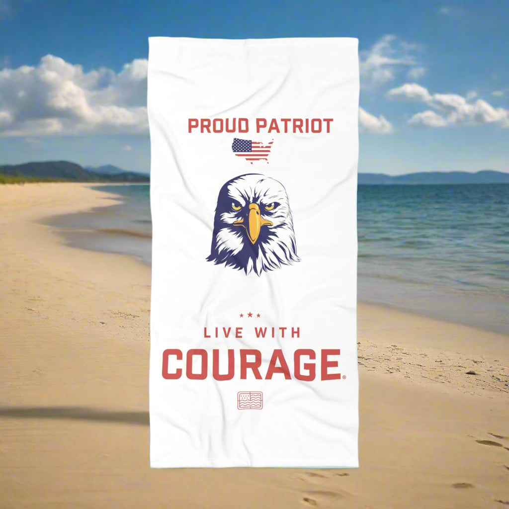 A beach towel in front of a beach scene with the American Eagle design and the text 'Proud Patriot' and the Live with Courage®️ logo.