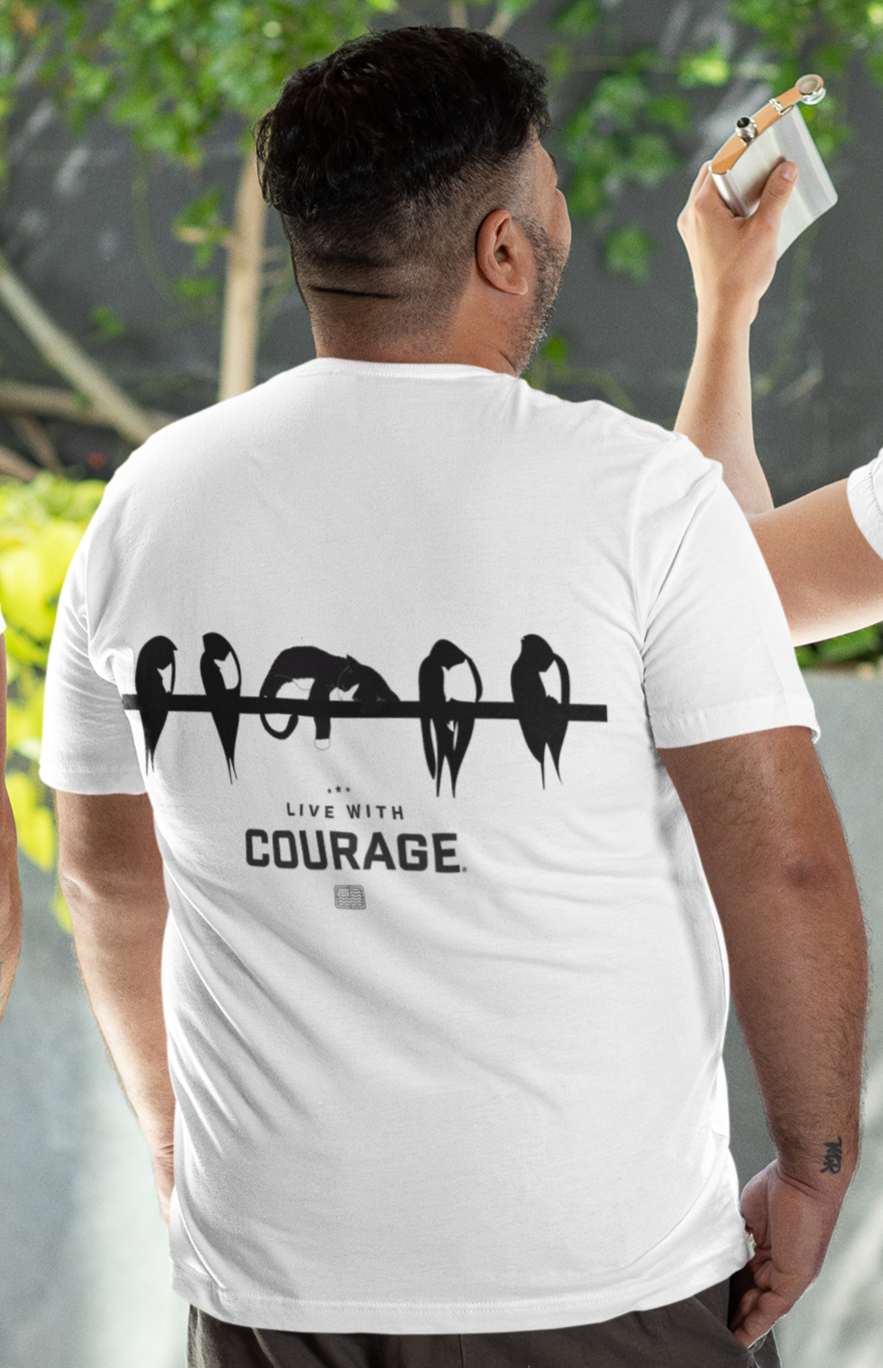 Person wearing a white t-shirt with 'Live with Courage' text on the back, standing outdoors with a graphic of a cat lying on a telephone wire along with birds.