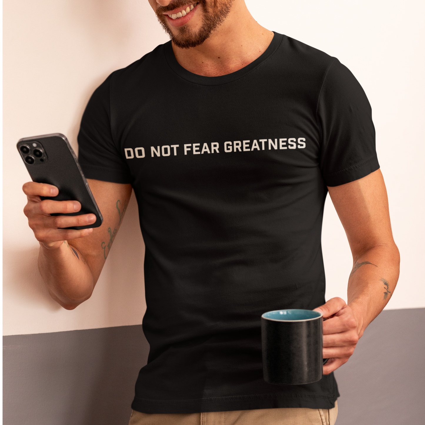 Person wearing a black t-shirt with 'DO NOT FEAR GREATNESS' text, holding a phone and cup.