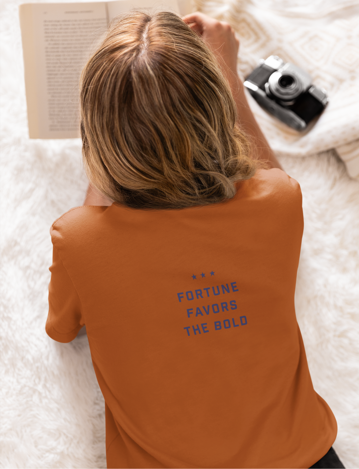 Person wearing an orange shirt with 'Fortune Favors the Bold' text, lying on a white surface with a book and camera.