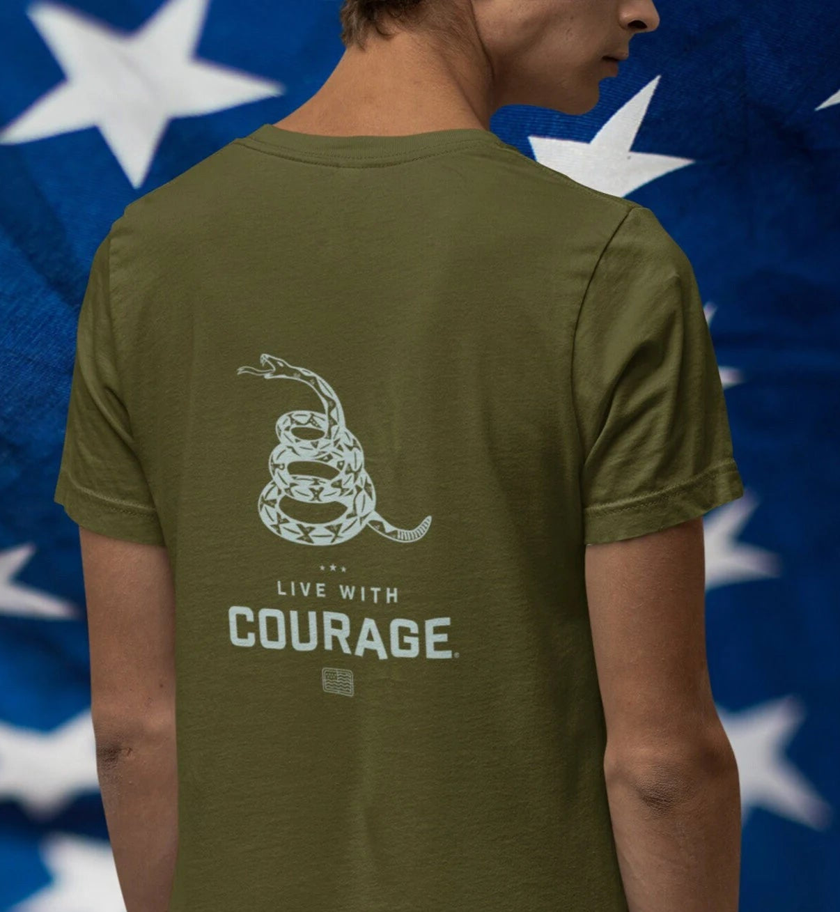 A person wearing a green short-sleeved t-shirt with a graphic of a coiled rattlesnake and the text 'LIVE WITH COURAGE' on the back, against a background of the American flag.