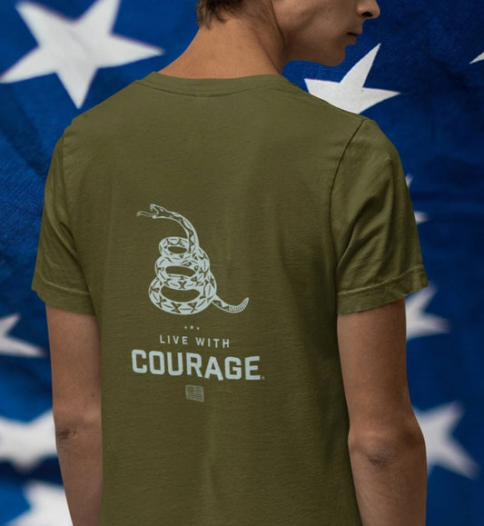 A person wearing a green short-sleeved t-shirt with a graphic of a coiled rattlesnake and the text 'LIVE WITH COURAGE' on the back, against a background of the American flag.
