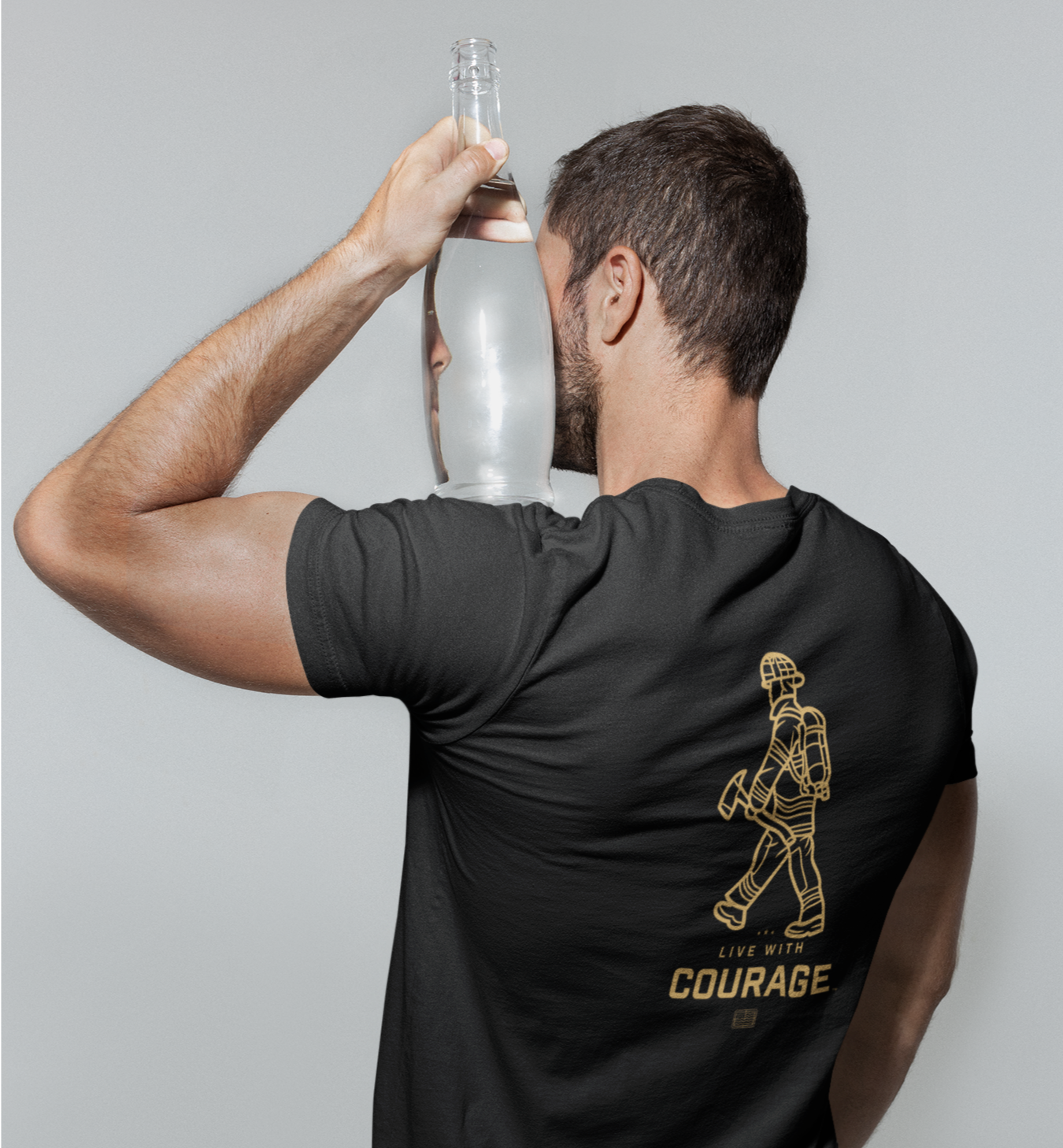 A person wearing a black unisex short sleeve tee holding a water bottle with a graphic of a firefighter and the words ' LIVE WITH COURAGE' on the back.