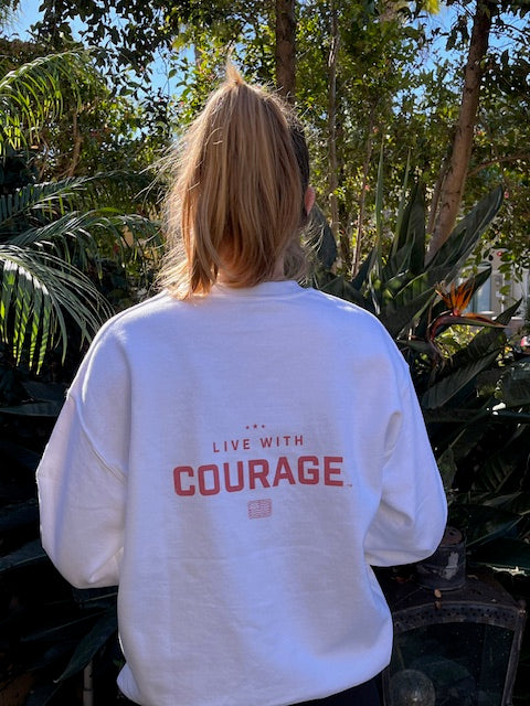 A woman facing some trees with a white sweatshirt and the words Live With Courage.