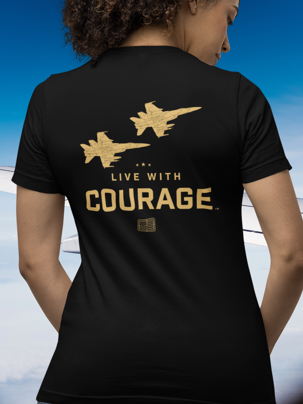 A person wearing a black short sleeve tee with a graphic of two fighter jets and the text 'LIVE WITH COURAGE' on the back.