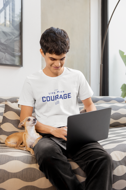 A youth wearing a white short sleeve tee with the text 'Live With COURAGE' on the front, sitting on a couch with a laptop and a dog beside him.