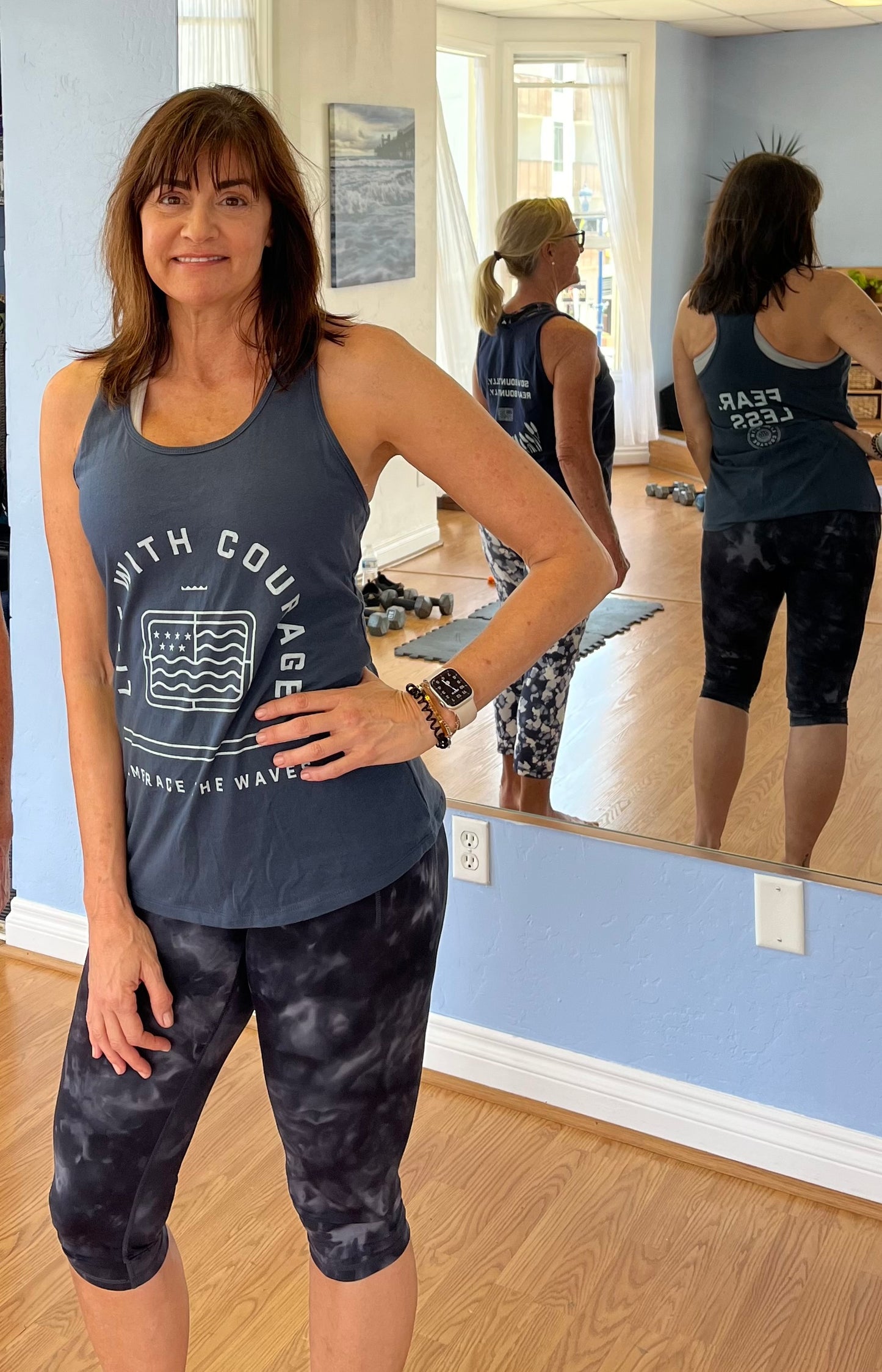 A woman wearing a gray racerback tank top with the text 'FEAR LESS' on the back, standing in a room with a mirror.