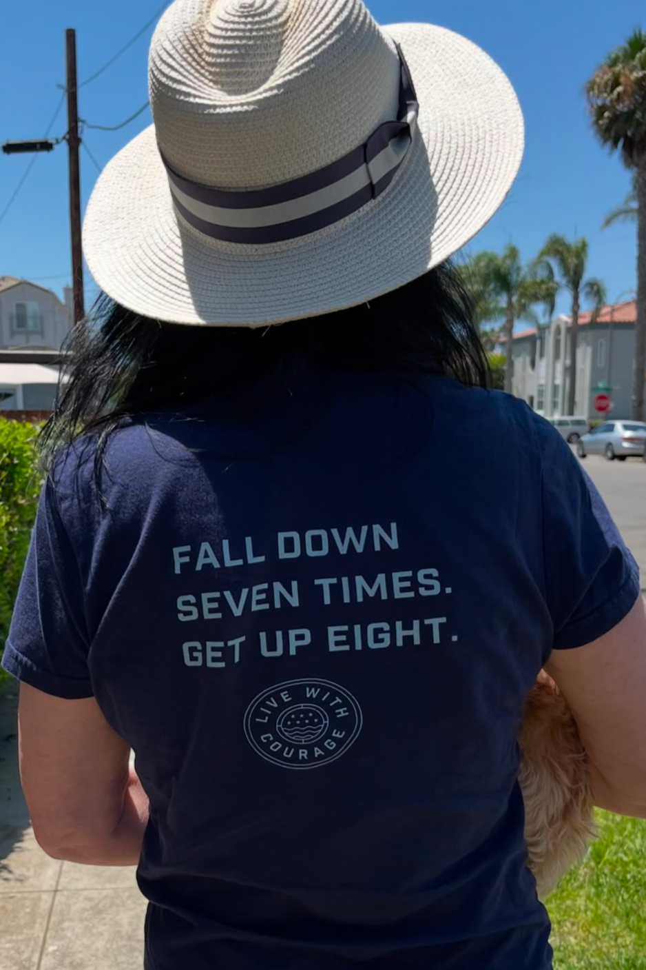 A woman wearing a navy blue crew neck t-shirt with the phrase 'FALL DOWN SEVEN TIMES, GET UP EIGHT' printed on the back. She is also wearing a wide-brimmed hat.