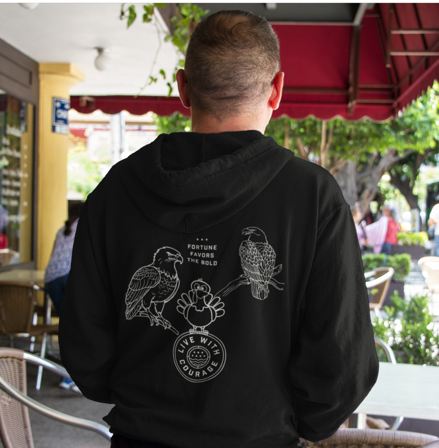 Person wearing a black hoodie with a a picture of two eagles on branches and a cartoon turkey atop the Live With Courage®️logo and the words Fortune Favors the Bold. 