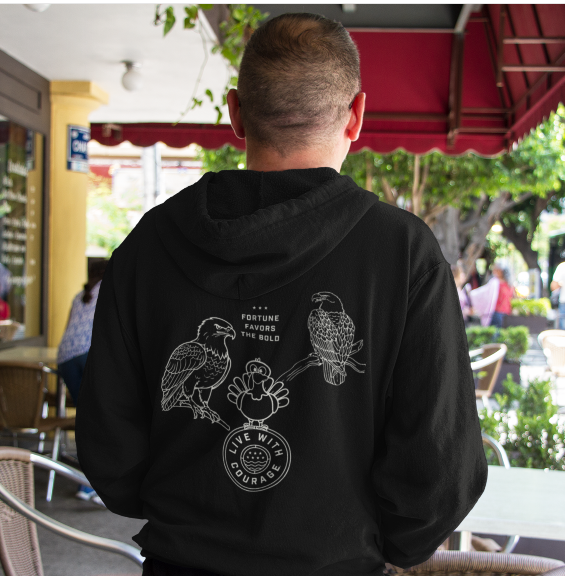 Person wearing a black hoodie with a a picture of two eagles on branches and a cartoon turkey atop the Live With Courage®️logo and the words Fortune Favors the Bold. 
