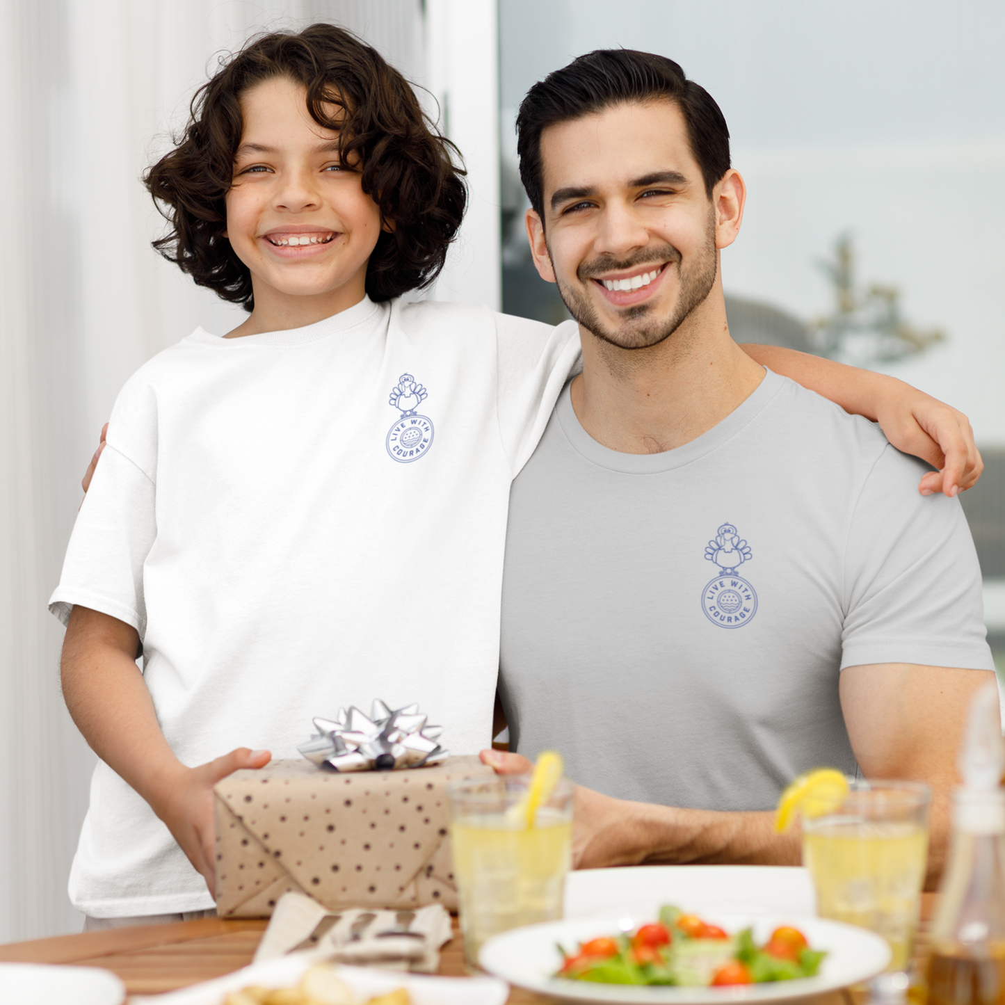 Father and son share a smile at the dinner table while wearing Live With Courage® Thanksgiving t-shirts with a turkey design on the front left chest