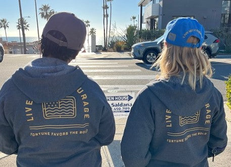 Two women wearing grey zip hoodies with the Live With Courage logo on the back facing the ocean.