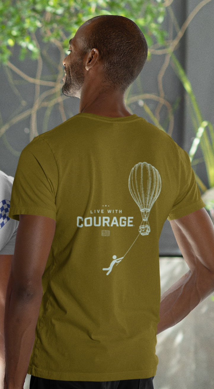 Person wearing a green t-shirt with 'Live with Courage' design on the back and a drawing of a hot air balloon in the air with a cartoon person hanging on below holding a rope.