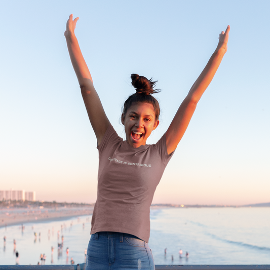 Woman jumping joyfully on a wooden pier with water and sky in the background with the word Courage is Contagious on the front of a grey t shirt