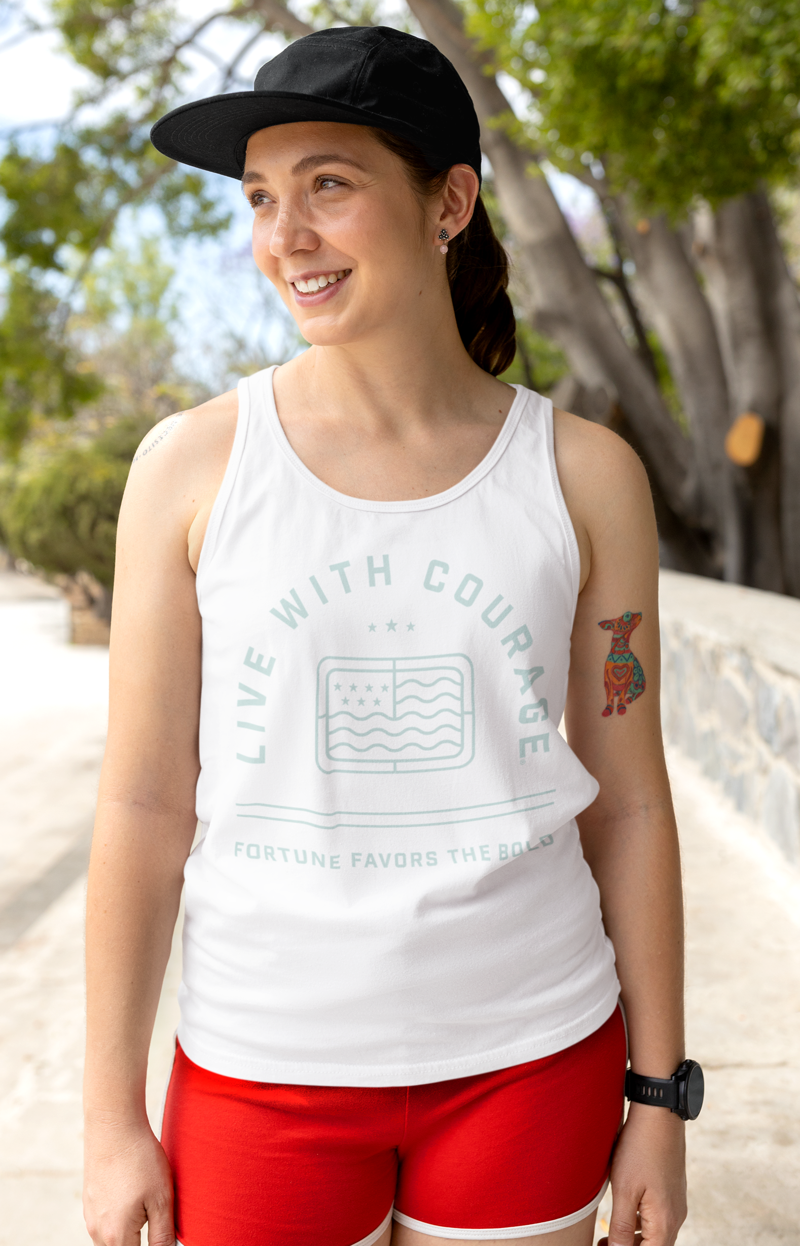 Woman wearing a white tank top with the words Live With Courage and Fortune Favors the Bold and red shorts standing outdoors on a path with trees.