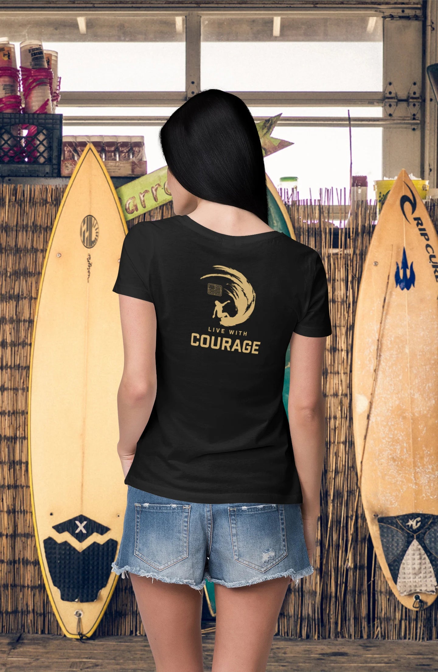 A woman facing away from the camera wears a black crew neck t-shirt with the text 'LIVE WITH COURAGE' and a surfing girl graphic on the back. She is standing in front of two surfboards.