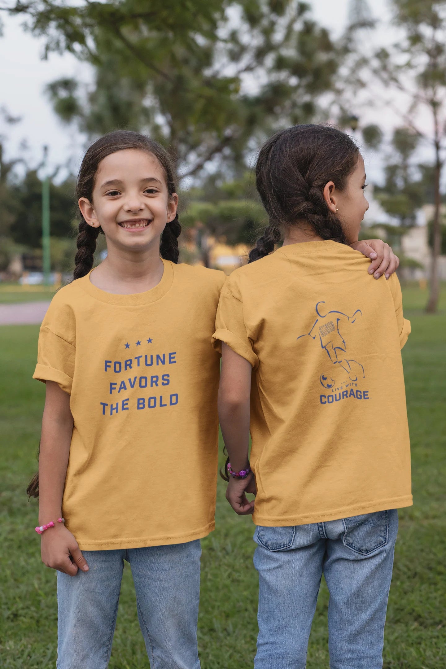 A pair of twin girls wearing yellow short sleeve t-shirts with text "Fortune Favors the Bold" and a soccer player graphic on them, standing outdoors. 