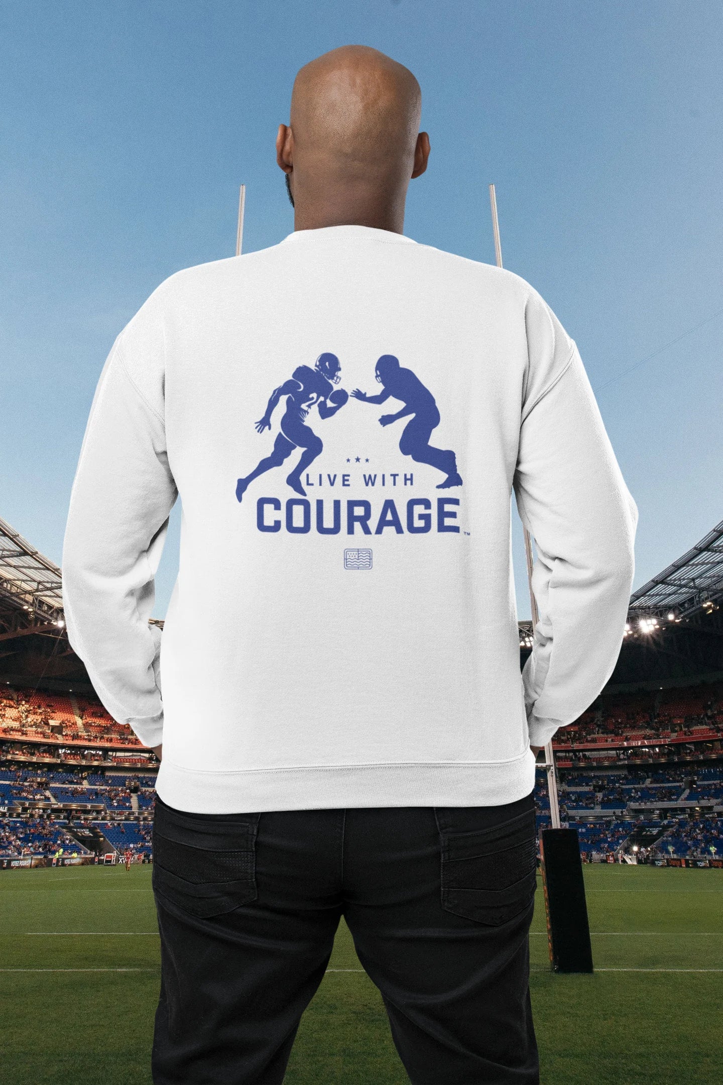 A person facing away from the camera wears a white crewneck sweatshirt with a logo and two blue figures playing football. Text on the logo reads 'Live With Courage.