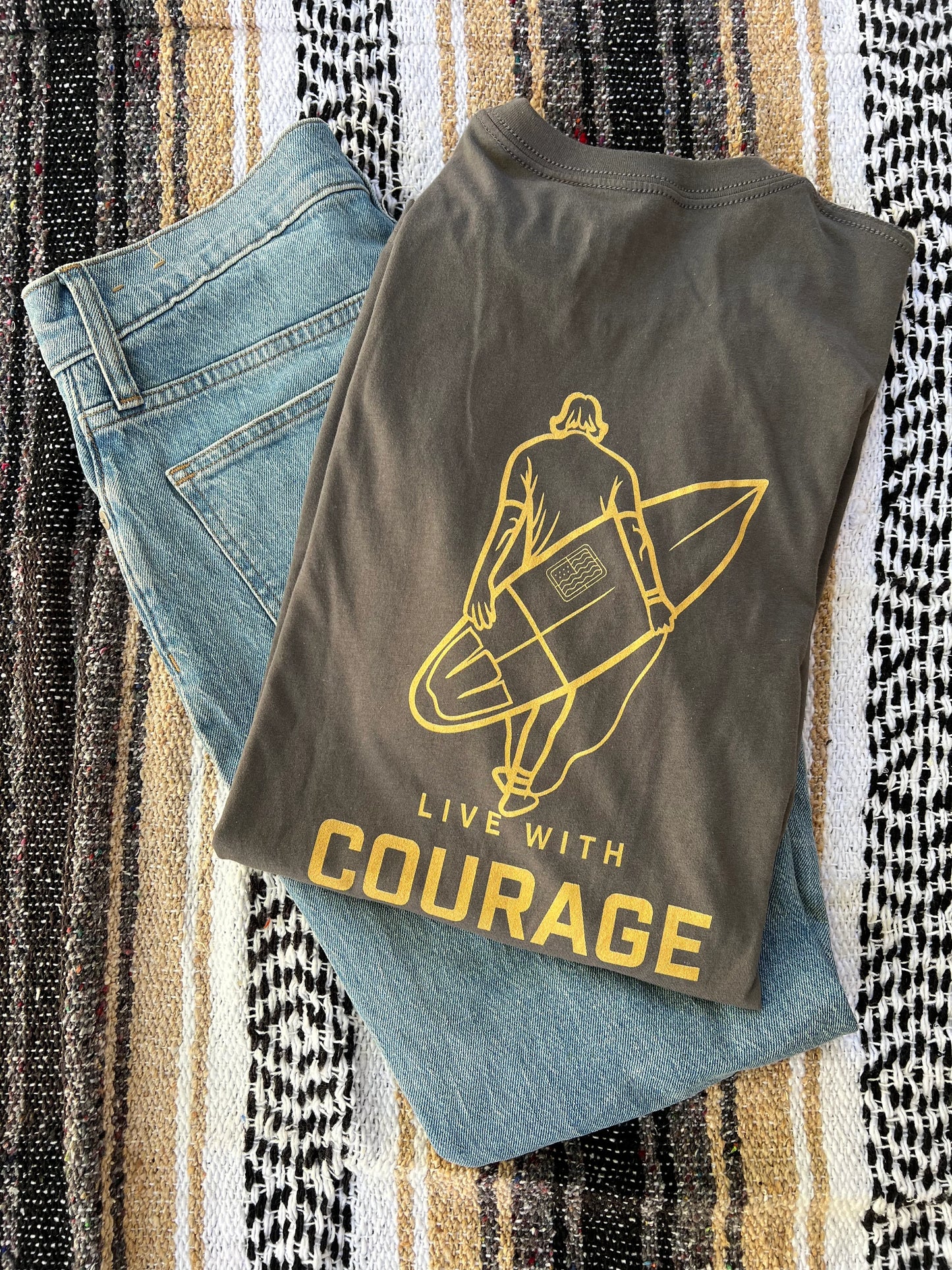 A gray unisex short sleeve tee with a surfboard design and the text 'LIVE WITH COURAGE' printed on the front, laid flat on a patterned surface.