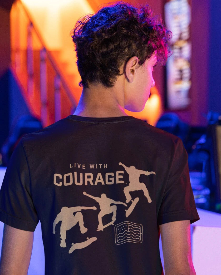 A person wearing a black short sleeve t-shirt with a graphic design on the back, featuring skateboarders and the text 'LIVE WITH COURAGE'.