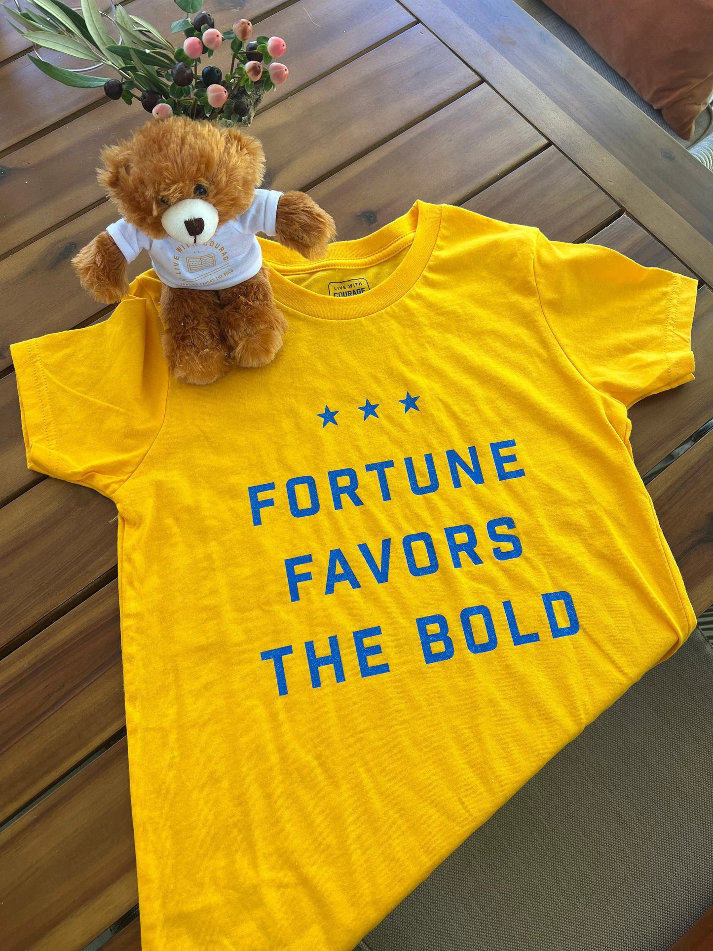 A yellow youth tee shirt laid out on a table with the words Fortune Favors the Bold and a small brown teddy bear.