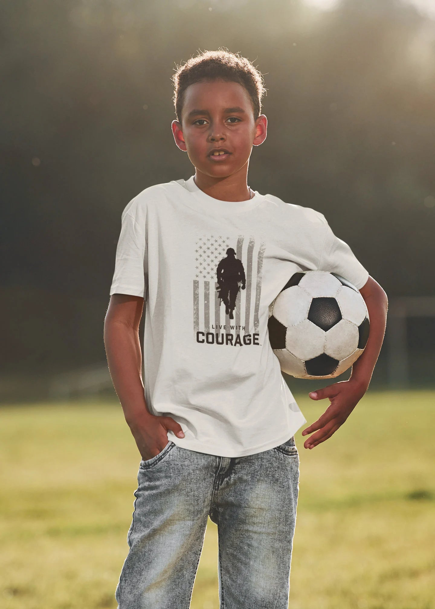 A youth wearing a white short-sleeve t-shirt with a graphic that includes the American flag and the silhouette of a soldier and the text 'LIVE WITH COURAGE', holding a soccer ball.