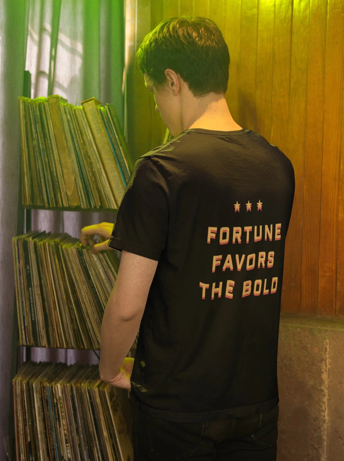 A person wearing a black t-shirt with 'Fortune Favors the Bold' printed on the back, standing in front of a shelf filled with vinyl records.