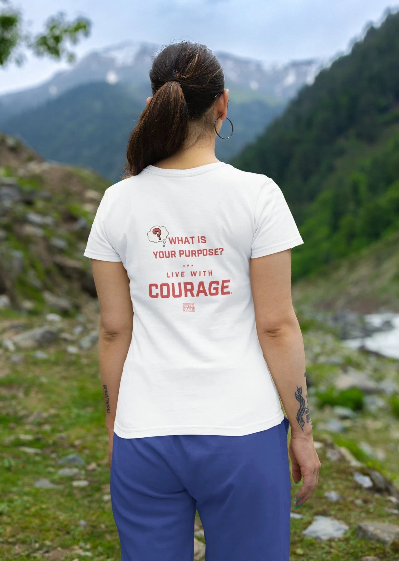 A woman facing away from the camera wears a white crew neck t-shirt with the text 'What is Your Purpose? Live with COURAGE' on the back, set against a mountainous landscape.