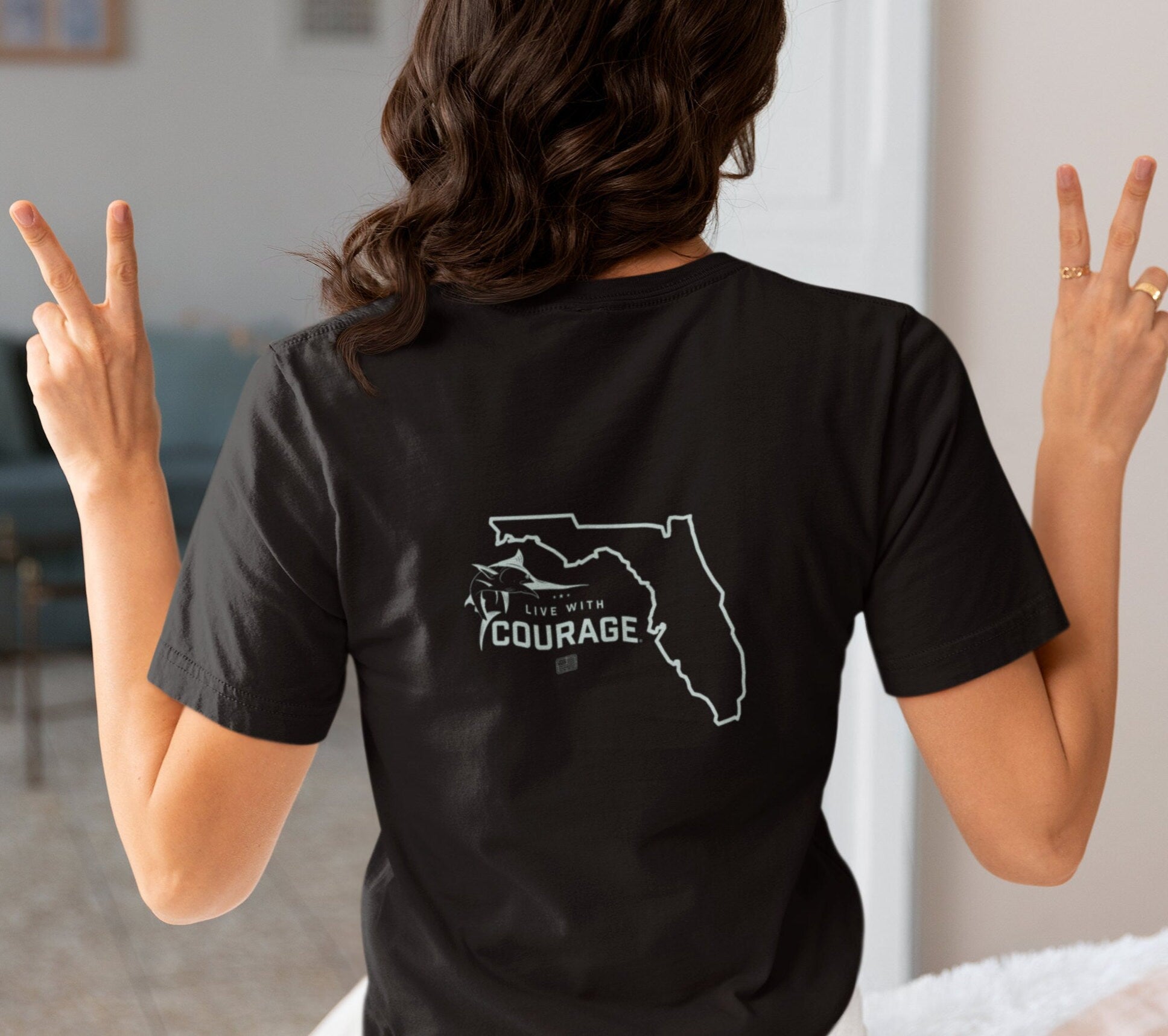 A woman wearing a black short sleeve tee shirt with an outline of the state of Florida and a graphic of a marlin jumping over the words Live With Courage®.