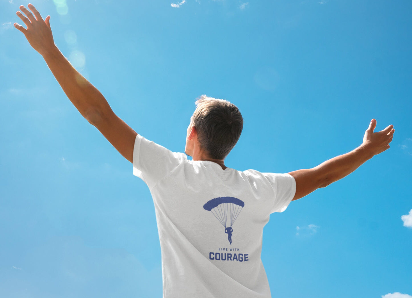 A man wearing a white unisex t-shirt reaching up for the blue sky. the shirt has a picture of a paraglider descending to earth and the words Live With Courage®️ 