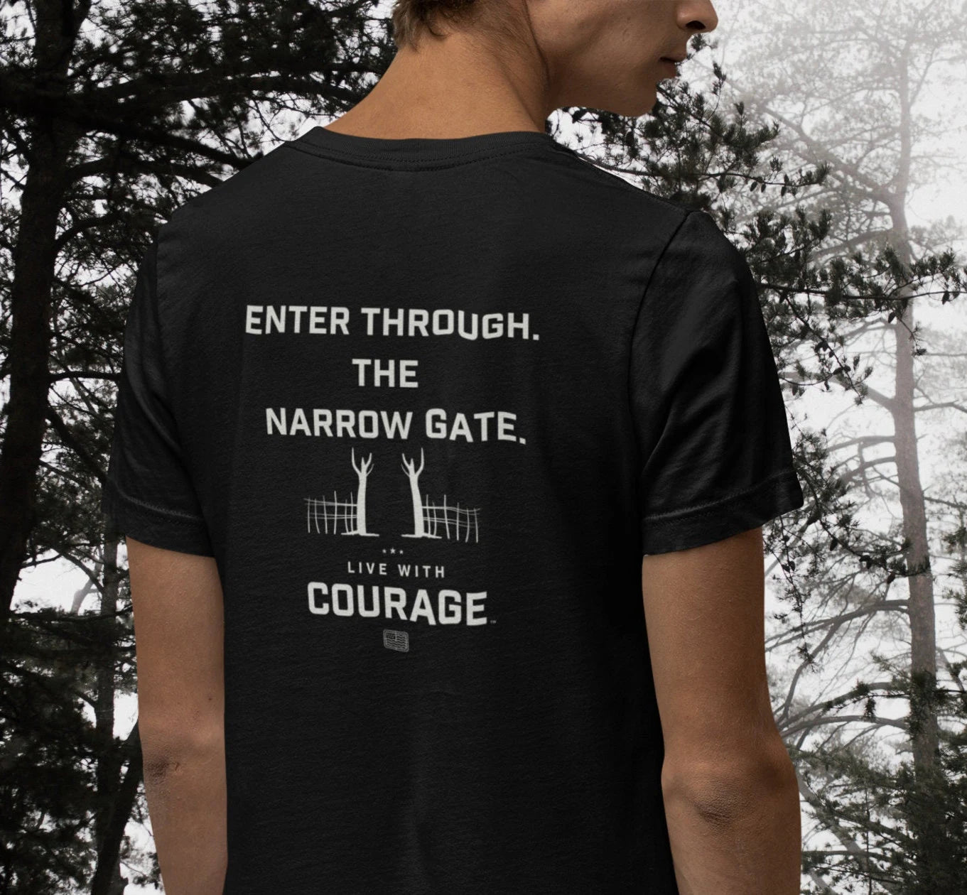 A person wearing a black unisex short sleeve tee with a quote from Matthew 7:13-14 Enter Through the Narrow Gate printed on the back.