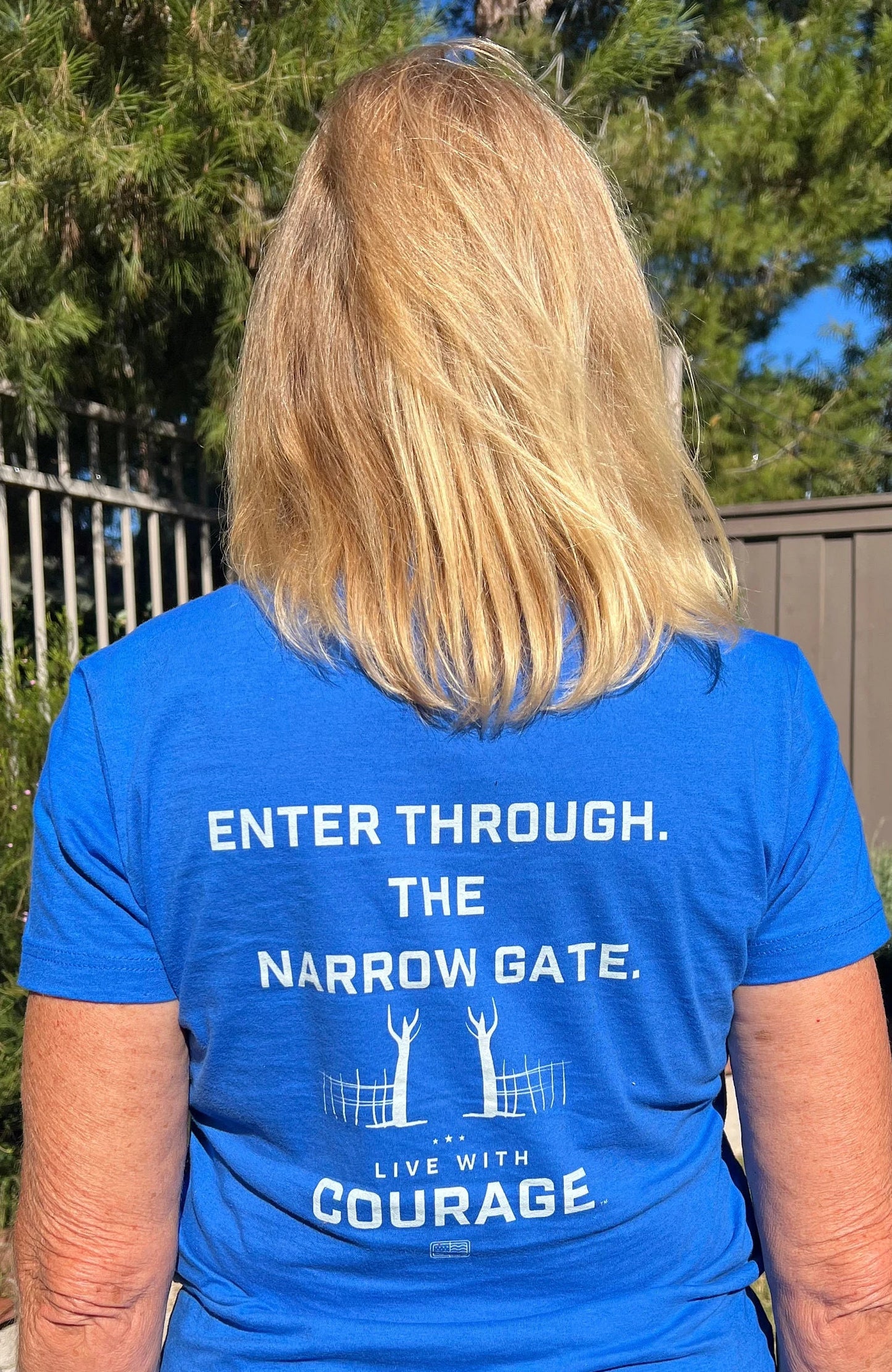 A woman wearing a blue crew neck t-shirt with the text 'ENTER THROUGH THE NARROW GATE. LIVE WITH COURAGE' printed on the back.