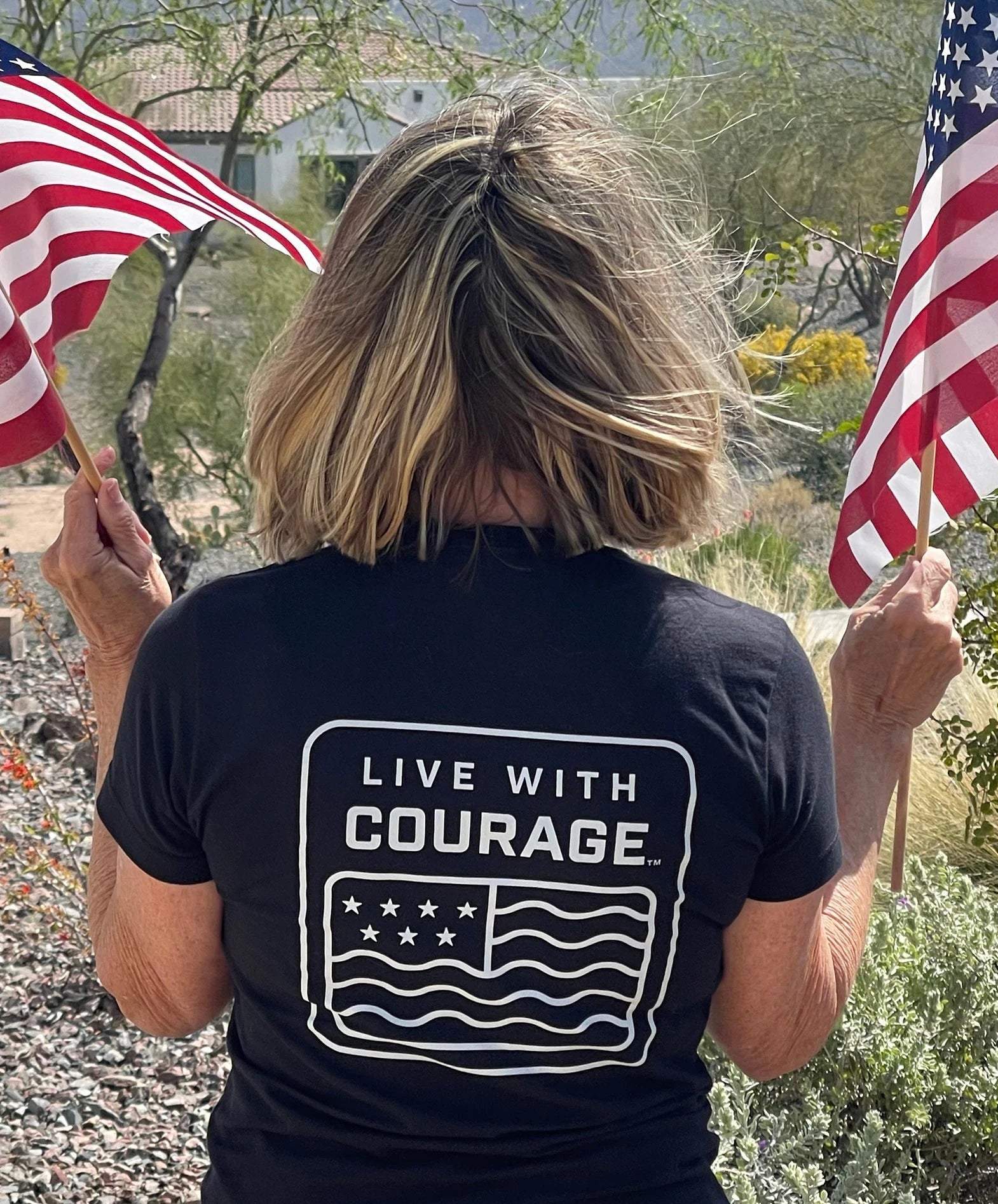 A woman facing away from the camera wears a black crew neck t-shirt with the Live With Courage stylized American flag design on the back. She is holding American flags in front of her.