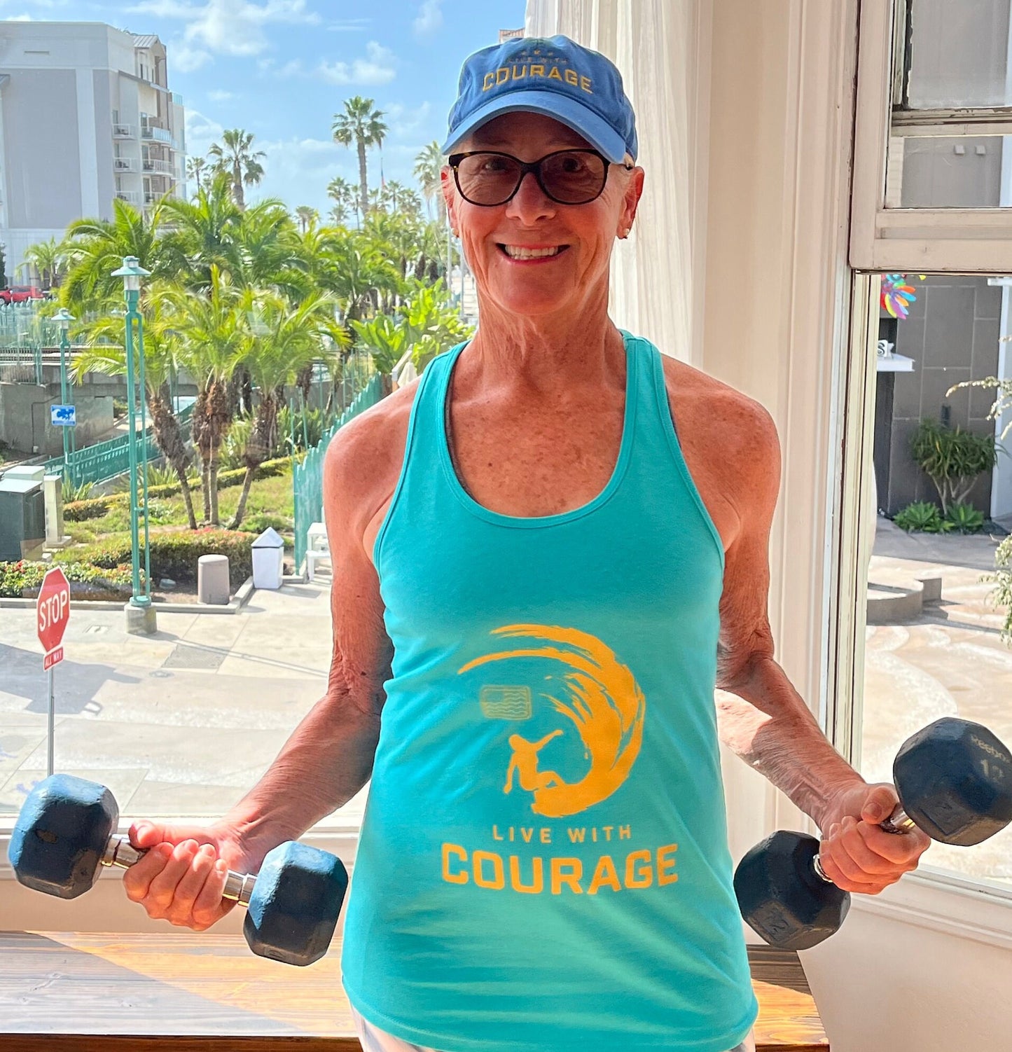 A woman wearing a turquoise racerback tank top with a logo that reads 'Live With Courage' and a graphic of a surfer girl inside a wave.