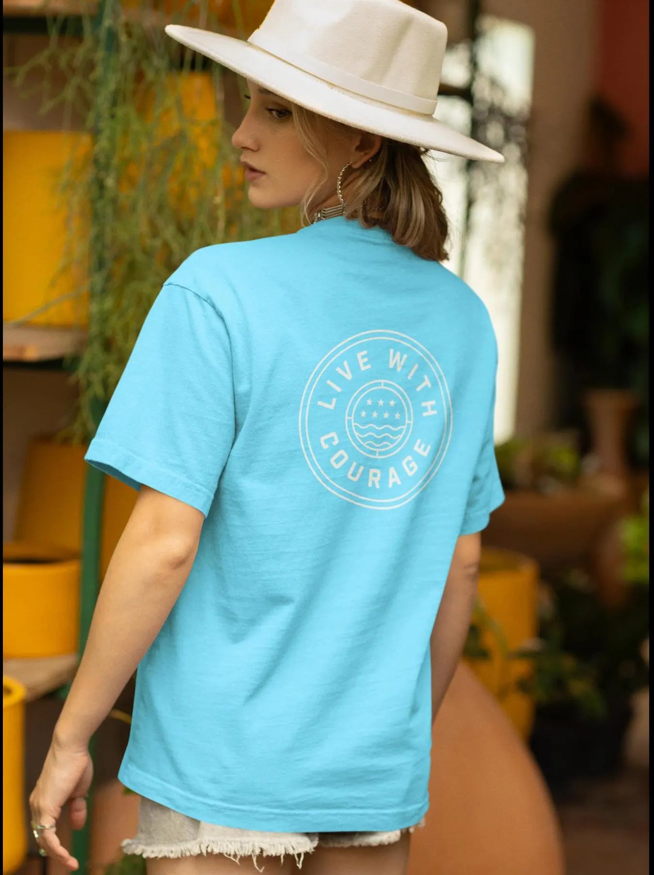 A person wearing a blue unisex short-sleeve t-shirt with a circular Live With Courage logo on the back, standing with their back towards the camera.