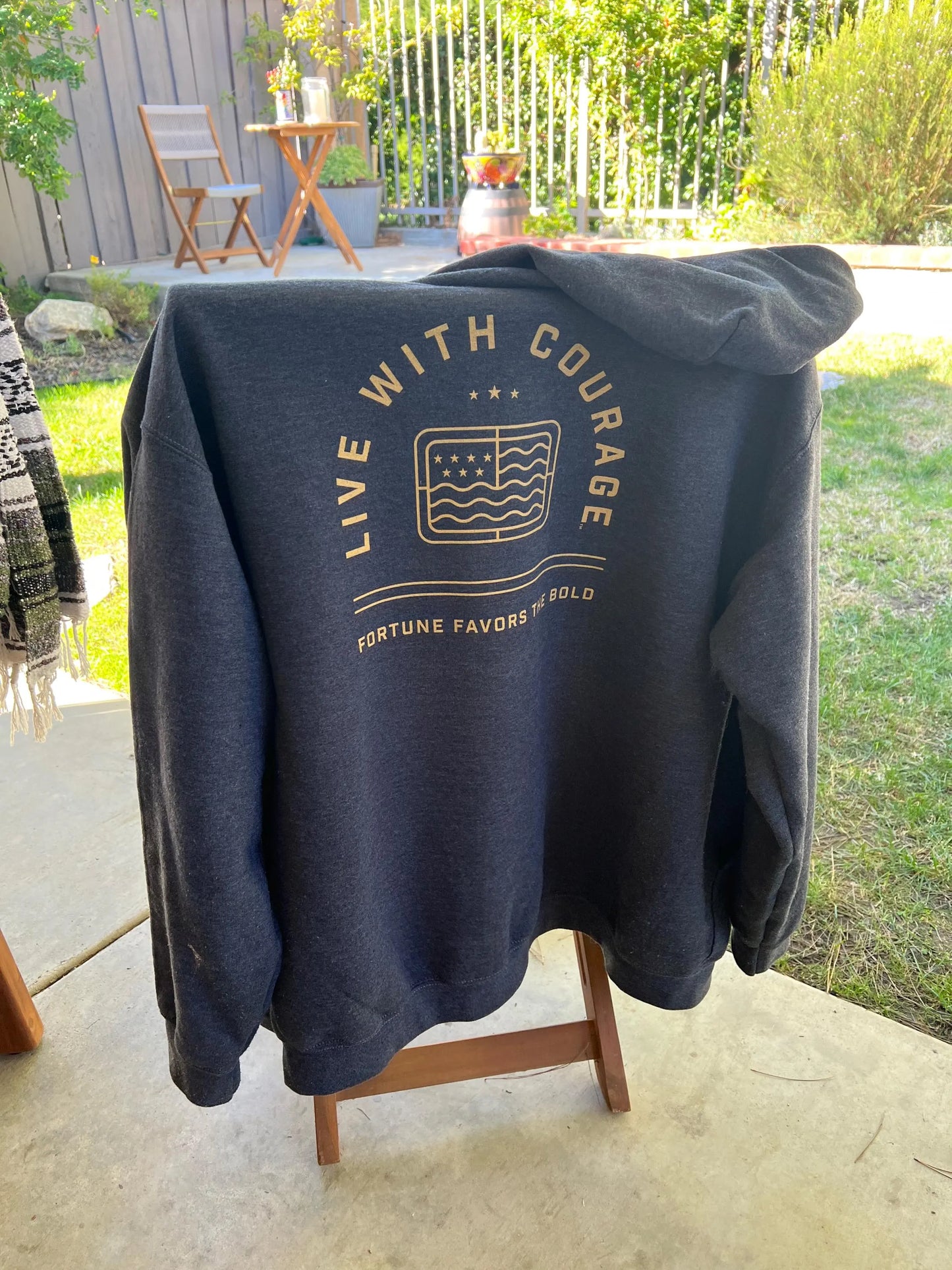 A grey full zip hoodie displayed on a chair outdoors, with the 'Live With Courage' logo printed on the back and the 'Fortune Favors the Bold' text on the front.