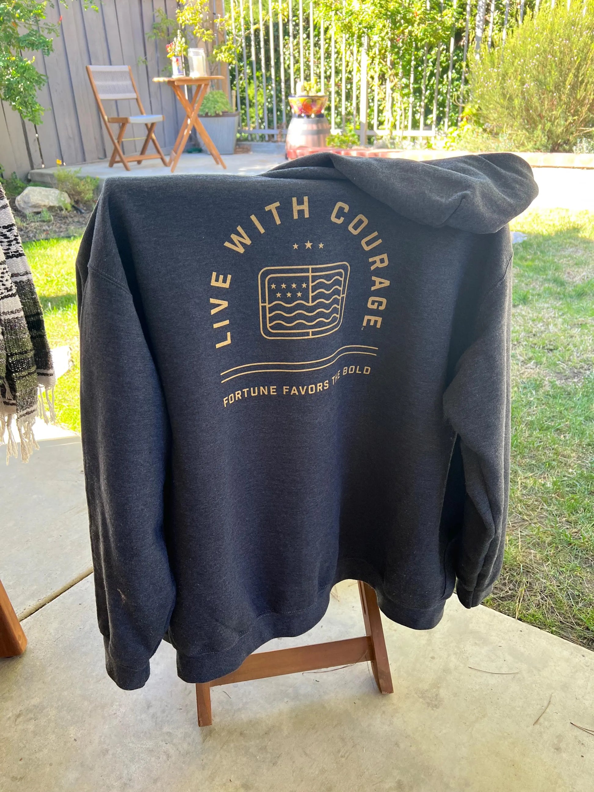 A grey full zip hoodie displayed on a chair outdoors, with the 'Live With Courage' logo printed on the back and the 'Fortune Favors the Bold' text on the front.