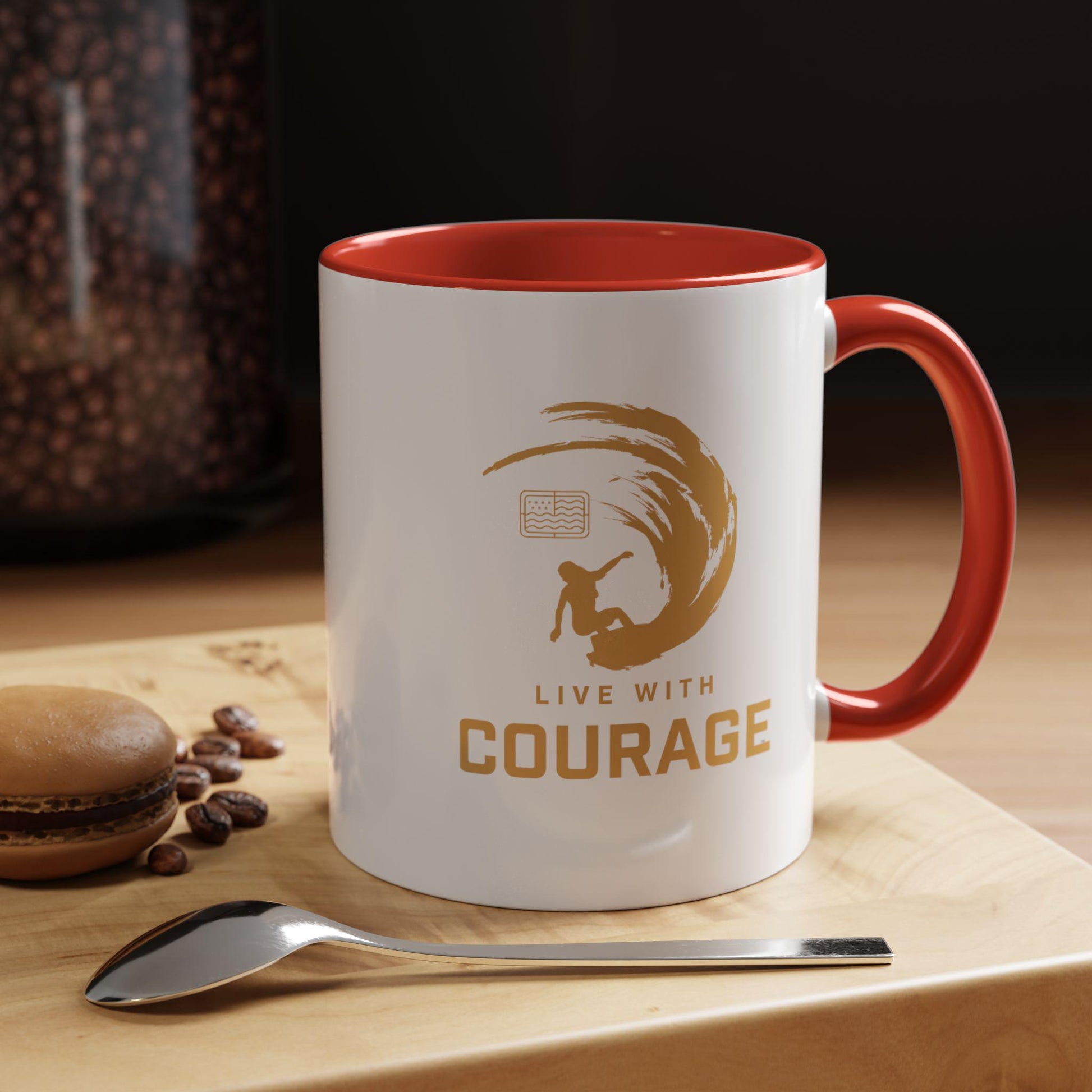 White ceramic coffee mug with red interior and handle featuring a surfer silhouette and the text 'LIVE WITH COURAGE', along with the logo 'Embrace the Waves'. The mug is displayed on a wooden surface with coffee beans and a spoon in the background.