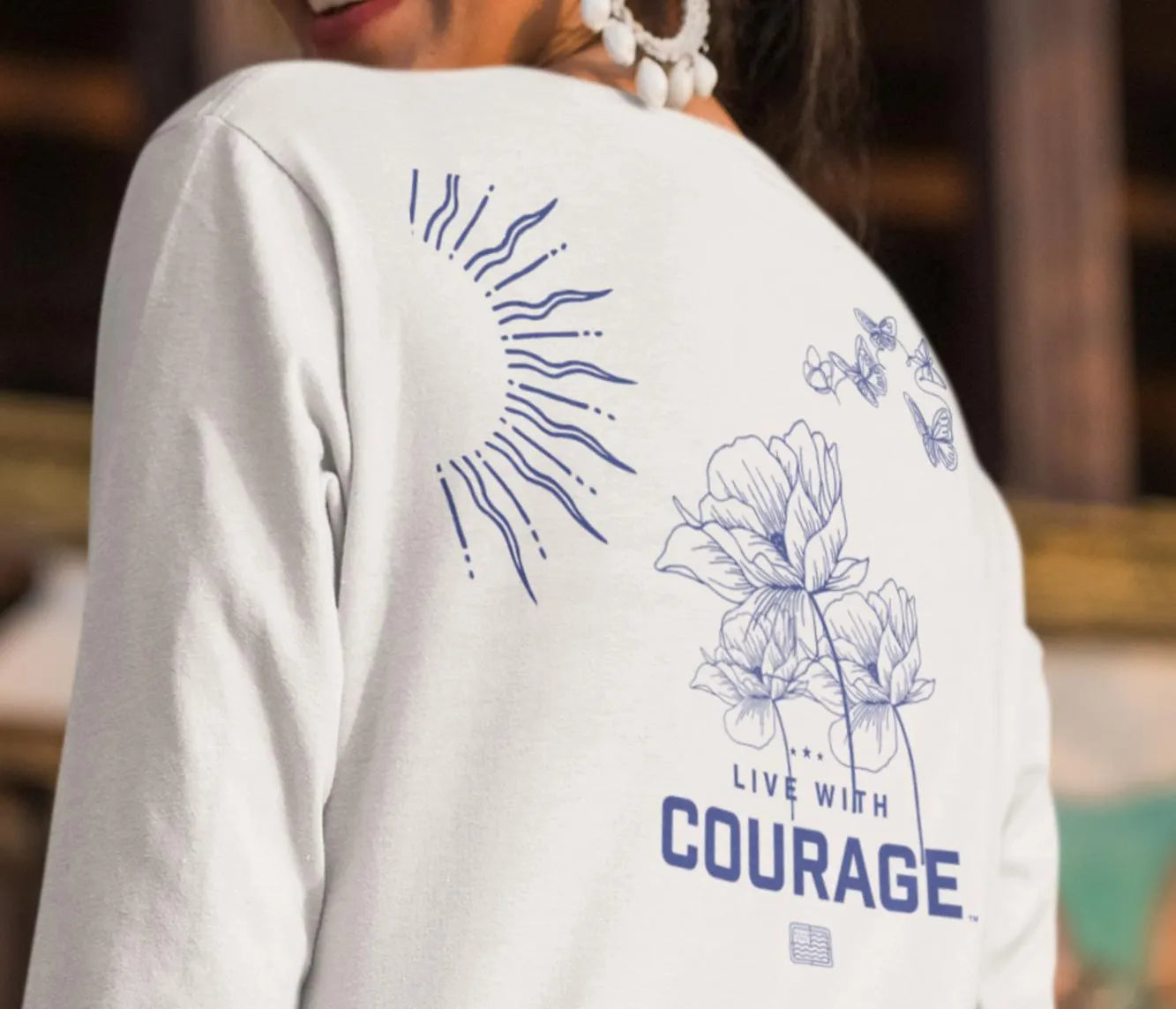 A person wearing a long sleeve white t-shirt with a floral and butterflies design, and text that reads 'LIVE WITH COURAGE'.