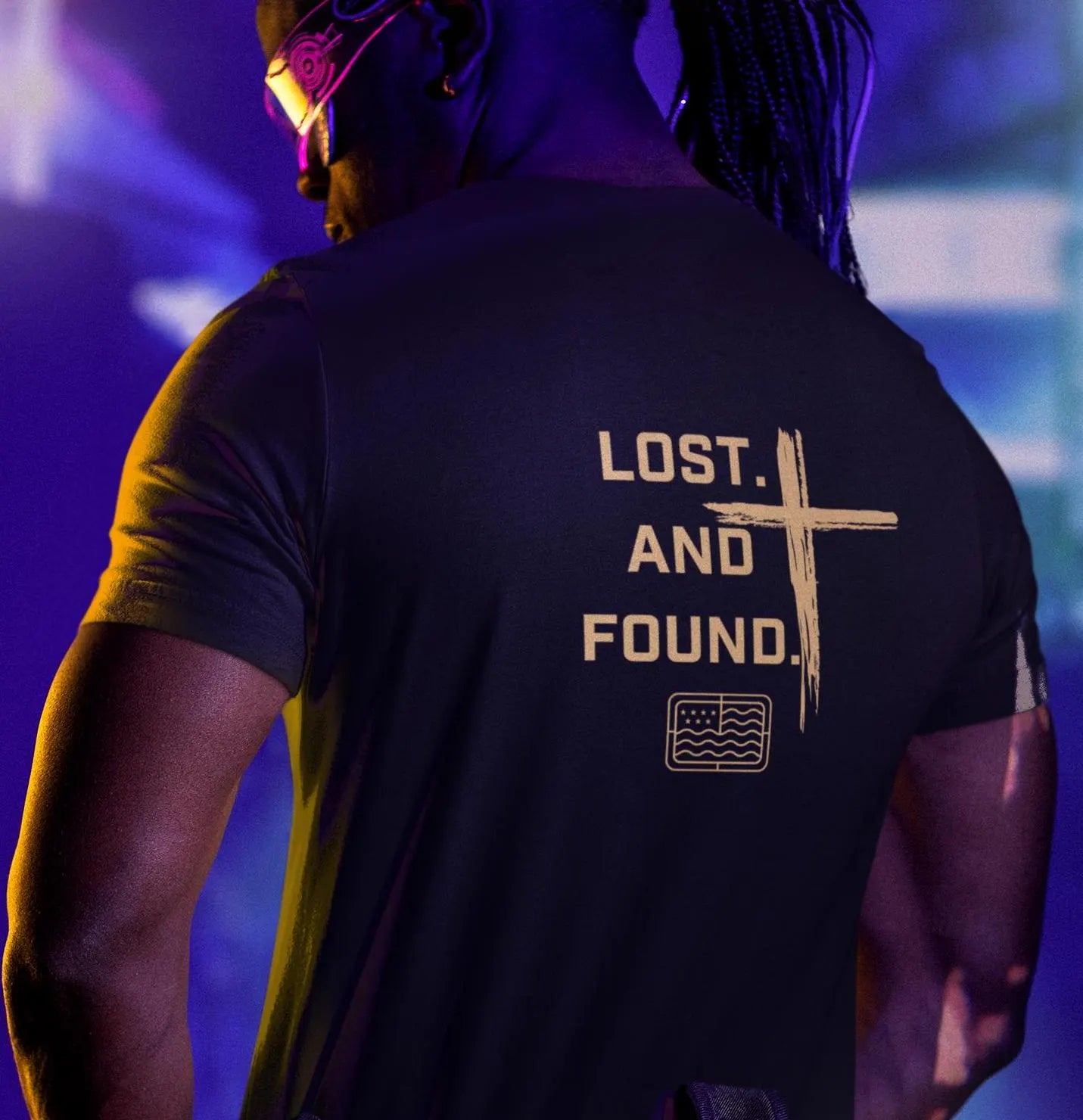 A person wearing a black unisex short sleeve tee with the text 'LOST. AND FOUND.' and a cross graphic on the back.