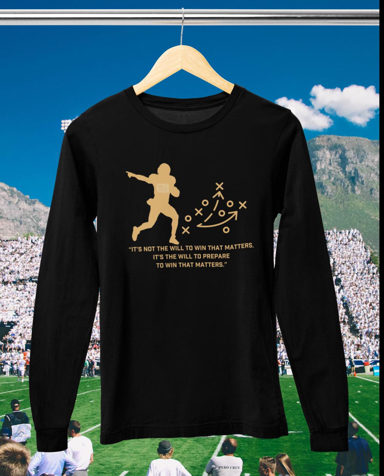 A black long sleeve tee with a graphic of a football player and a quote from Bear Bryant on the front, hanging on a display against a football stadium backdrop.