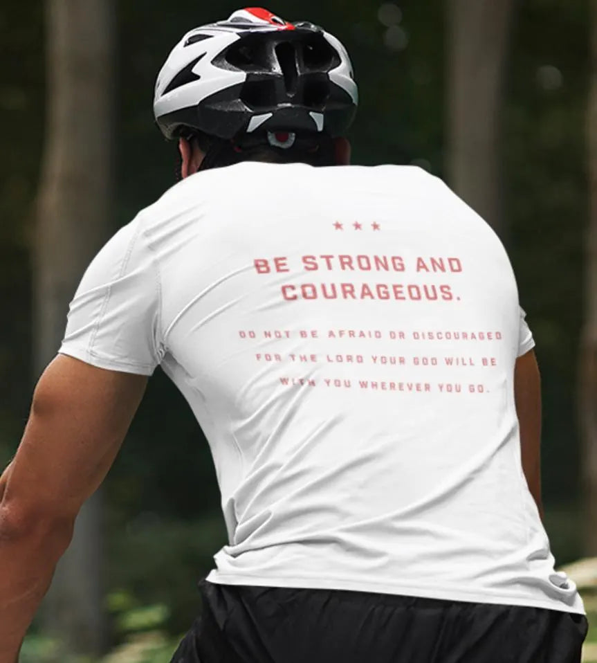 A person riding a bicycle wearing a white crew neck t-shirt with the back text 'BE STRONG AND COURAGEOUS. Do not be afraid or discouraged, for the Lord your God will be with you wherever you go.