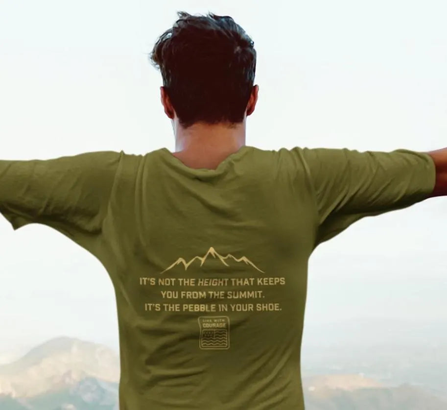 A person wearing a green short-sleeve t-shirt with a mountain range design and text on the back that says "it's not the height that keeps you from the summit. It's the pebble in your shoe. And the words Live With Courage.