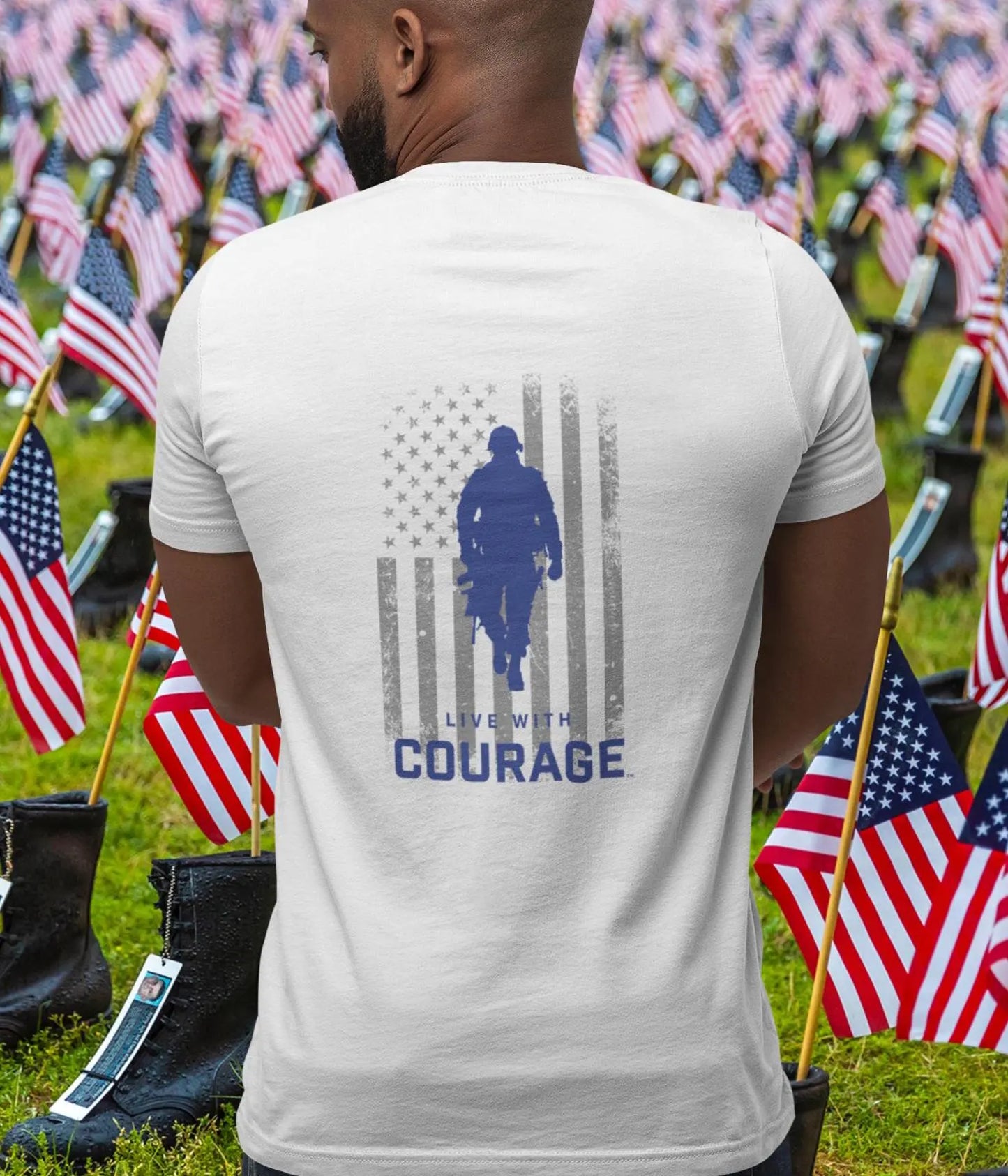 A person wearing a white crew neck t-shirt with a silhouette of a soldier and the flag of the United States on the back, along with the text 'LIVE WITH COURAGE'. The t-shirt is showcased in a field of American flags.
