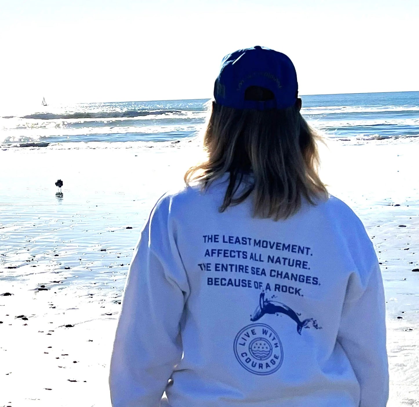 A person standing on a beach facing away from the camera wearing a white sweatshirt with a quote from mathematician Blaise Pasqual and a Live With Courage logo on the back, and a cap on the head.