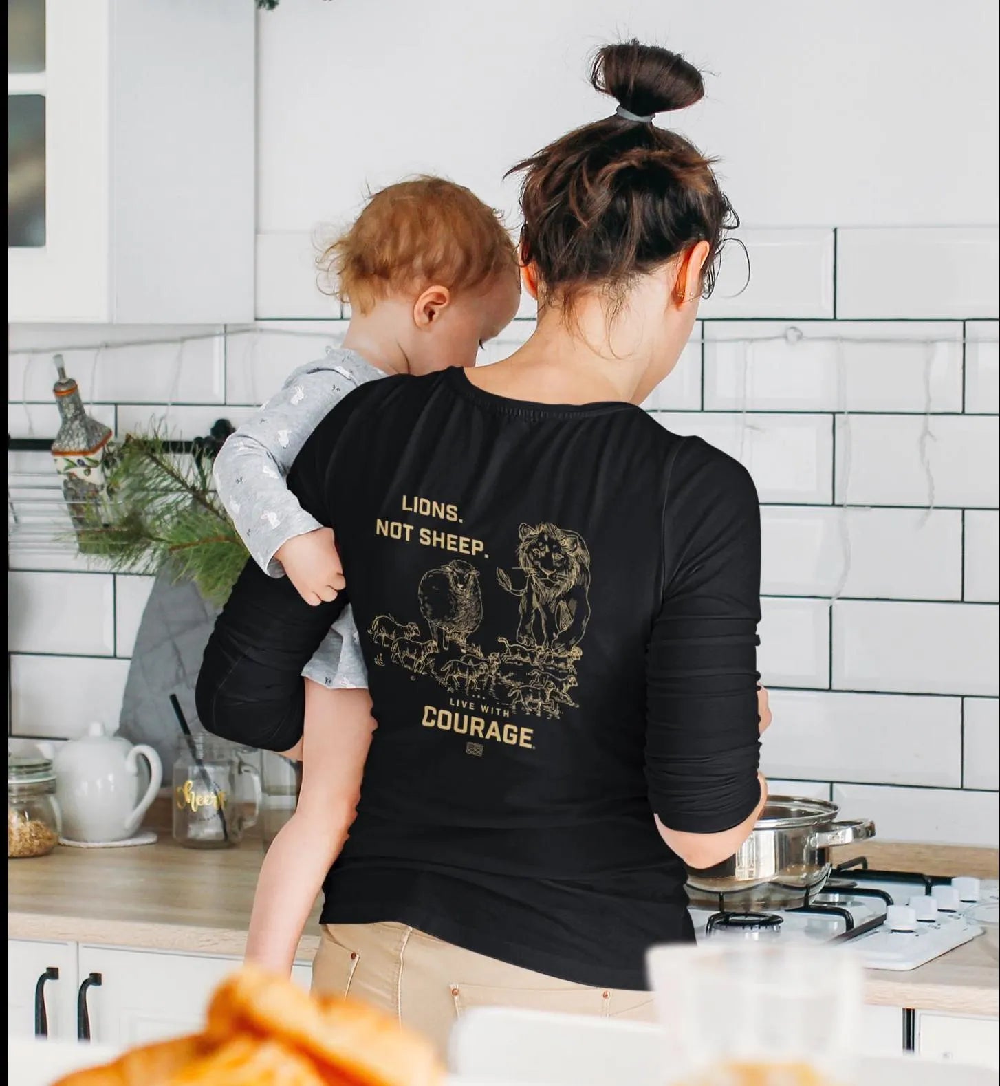 A person wearing a black long sleeve tee with the words Lions Not Sheep and a lion graphic on the back, standing in a kitchen holding a baby.