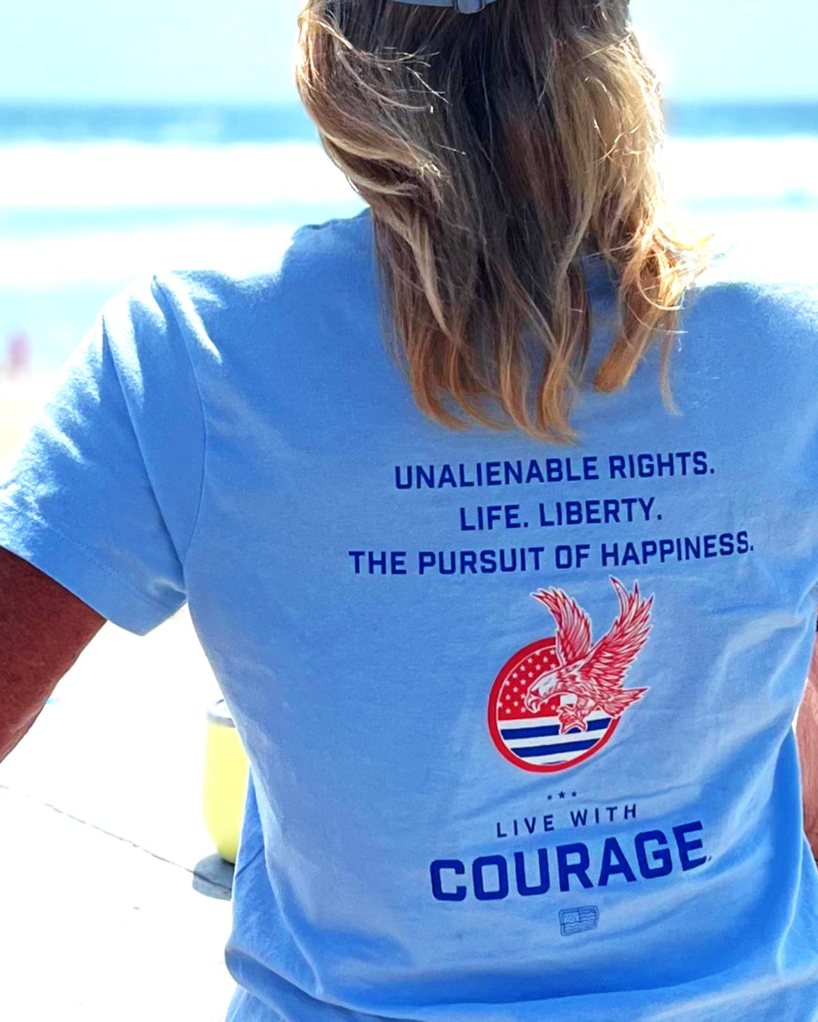 Person wearing a blue t-shirt with a patriotic design and text "Unalienable Rights. Life. Liberty. The Pursuit of Happiness and Live With Courage" on the beach.