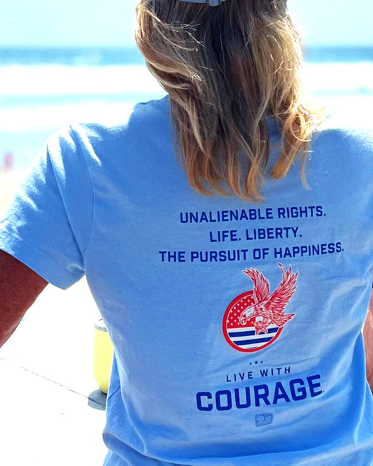 Person wearing a blue t-shirt with a patriotic design and text "Unalienable Rights. Life. Liberty. The Pursuit of Happiness and Live With Courage" on the beach.